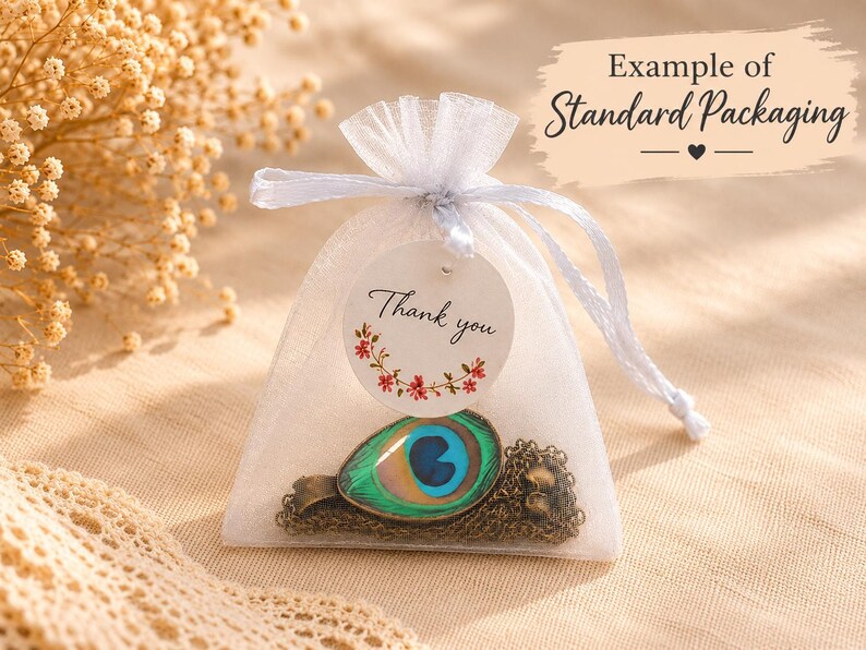 Puede incluir: Una bolsa de organza blanca con cord&oacute;n que contiene un collar con un colgante de ojo de pluma de pavo real. Una etiqueta redonda blanca unida al cord&oacute;n presenta el texto Thank you y un peque&ntilde;o gr&aacute;fico de corona floral en rojo y verde.