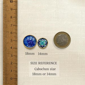 May include: Two round cabochons with gold-tone rims, one featuring a blue floral pattern and the other a blue and white geometric design. A wooden ruler shows the 18mm and 14mm sizes, alongside a one-euro coin for scale. Text reads SIZE REFERENCE.