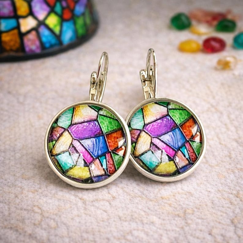 Puede incluir: Un par de pendientes con cierre de palanca que presentan un vibrante patr&oacute;n de vidrieras con segmentos geom&eacute;tricos en tonos morado, verde, azul, amarillo y naranja, enmarcados en un marco de metal plateado pulido.