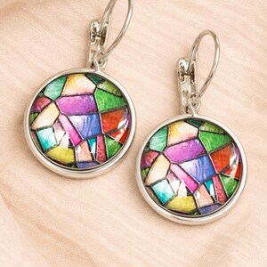 Puede incluir: Un par de pendientes colgantes de tono plateado con engastes circulares. Cada pendiente contiene un vibrante patr&oacute;n de mosaico multicolor con segmentos en verde, morado, azul, amarillo y rojo, bajo una cubierta de vidrio abovedada.