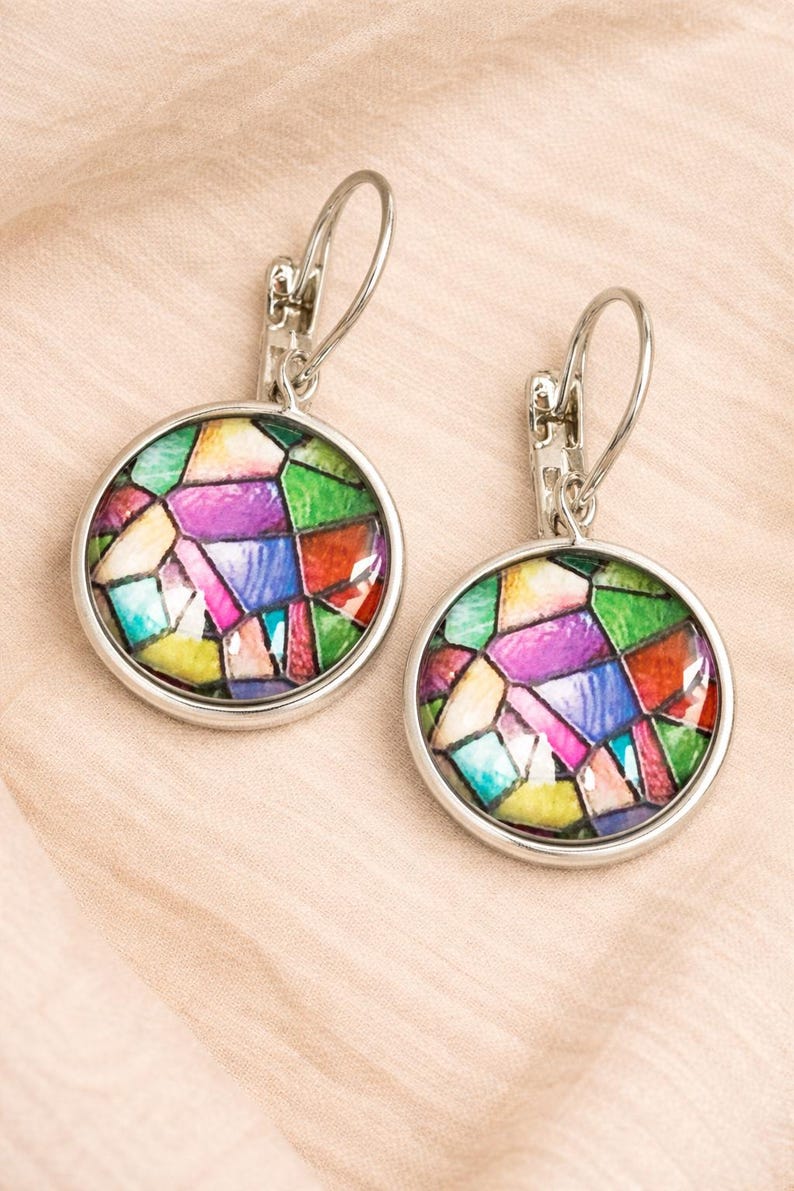 Puede incluir: Un par de pendientes colgantes de tono plateado con engastes circulares. Cada pendiente contiene un vibrante patr&oacute;n de mosaico multicolor con segmentos en verde, morado, azul, amarillo y rojo, bajo una cubierta de vidrio abovedada.