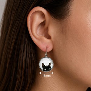 May include: A round silver-toned drop earring featuring a graphic illustration of a black cat's face against a white background. The circular pendant measures 18mm in diameter and hangs from a simple silver-toned hook wire attachment.