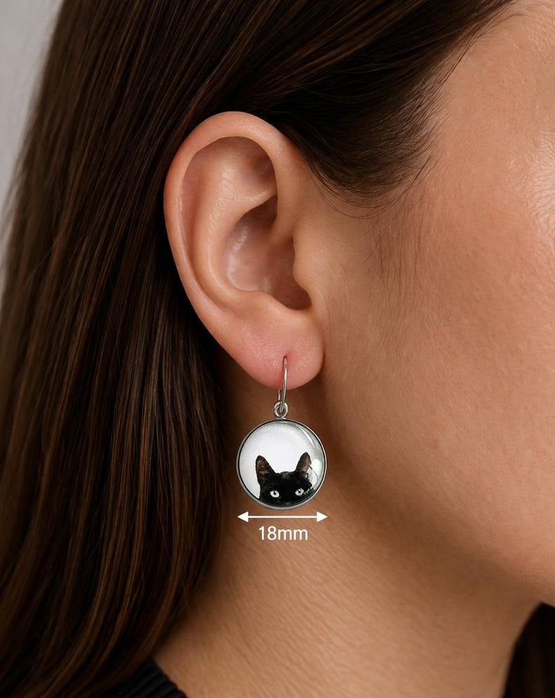 May include: A round silver-toned drop earring featuring a graphic illustration of a black cat's face against a white background. The circular pendant measures 18mm in diameter and hangs from a simple silver-toned hook wire attachment.