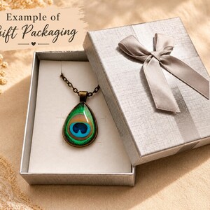 Puede incluir: Un collar con colgante en forma de l&aacute;grima que presenta un patr&oacute;n de pluma de pavo real en tonos verde, azul y dorado, suspendido de una cadena de metal oscuro, presentado en una caja de regalo plateada con un lazo a juego.