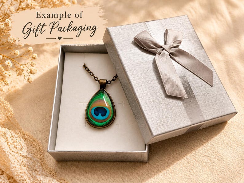 Puede incluir: Un collar con colgante en forma de l&aacute;grima que presenta un patr&oacute;n de pluma de pavo real en tonos verde, azul y dorado, suspendido de una cadena de metal oscuro, presentado en una caja de regalo plateada con un lazo a juego.