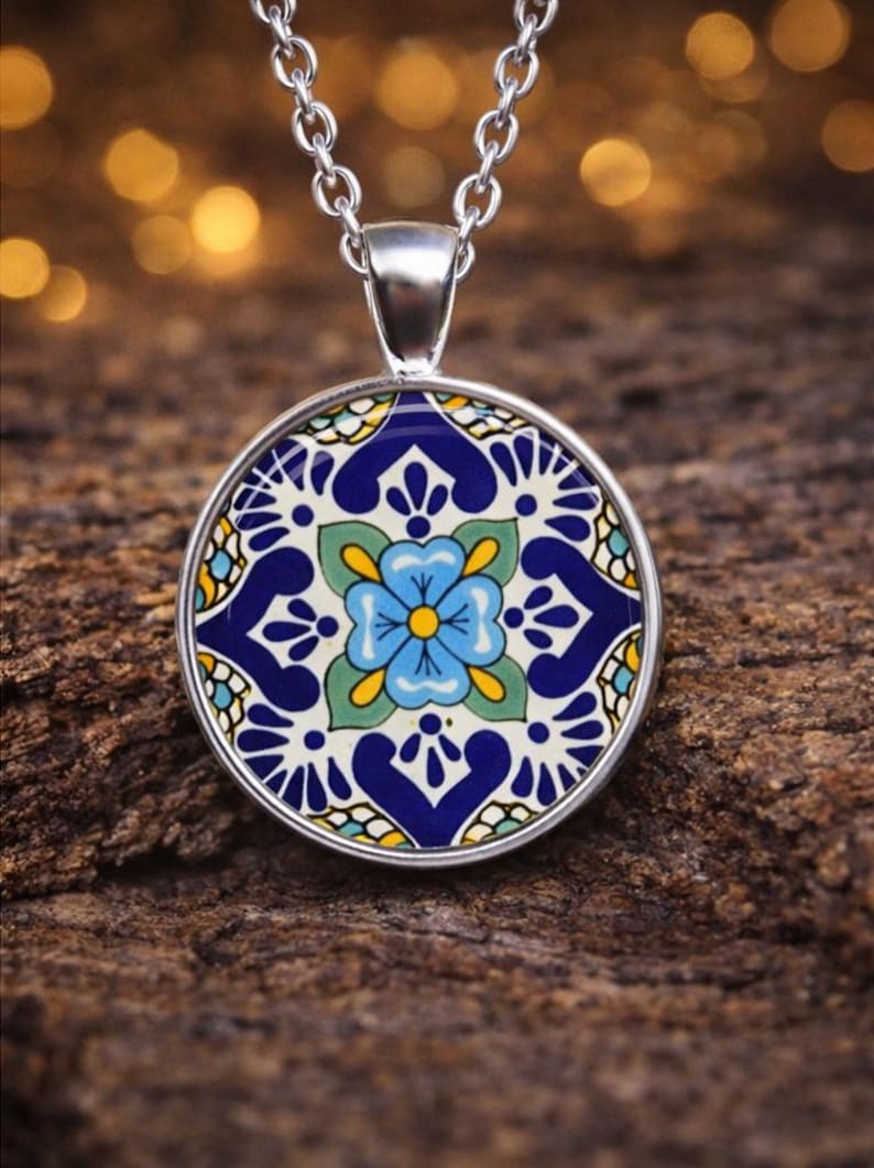 May include: A silver pendant necklace featuring a circular charm with a vibrant floral pattern in shades of blue, green, yellow, and white. The design displays a central light blue flower surrounded by intricate dark blue and yellow geometric motifs.