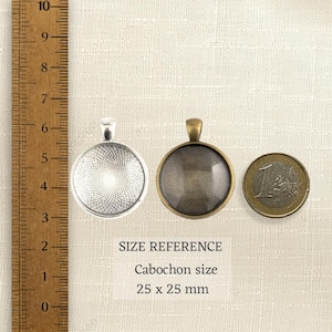 May include: Two round metal pendant trays with bails, one silver-toned with a textured pattern and one bronze-toned, positioned next to a wooden ruler and a one-euro coin. Text indicates a cabochon size of 25 x 25 mm for size reference.