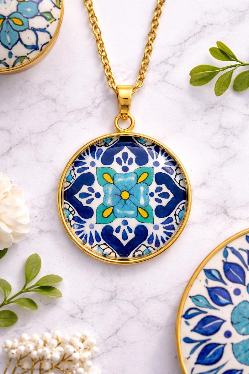 May include: A gold-toned pendant necklace with a round, patterned charm. The charm features a blue and white floral design with yellow accents. The necklace is displayed on a marble surface, with additional decorative elements.