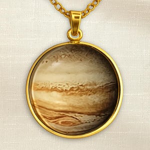 Puede incluir: Un collar con colgante dorado con un dije redondo que muestra una imagen de Júpiter. El dije tiene un bisel dorado y una cadena dorada. El planeta Júpiter se representa con patrones arremolinados marrones, beige y blancos.