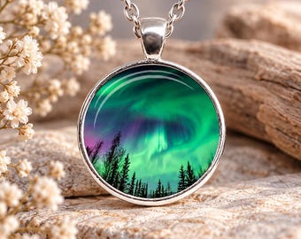 Puede incluir: Un collar con colgante circular de tono plateado que presenta una imagen vibrante de la aurora boreal con vetas de luz verde y púrpura sobre una silueta oscura de pinos. El colgante cuelga de una cadena de eslabones de metal plateado.