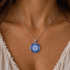 Puede incluir: Un collar de tono plateado con un colgante circular que presenta un patr&oacute;n floral en azul y blanco. El colgante cuelga de una delicada cadena de tono plateado que descansa sobre el pecho. El dise&ntilde;o muestra un motivo sim&eacute;trico y complejo.