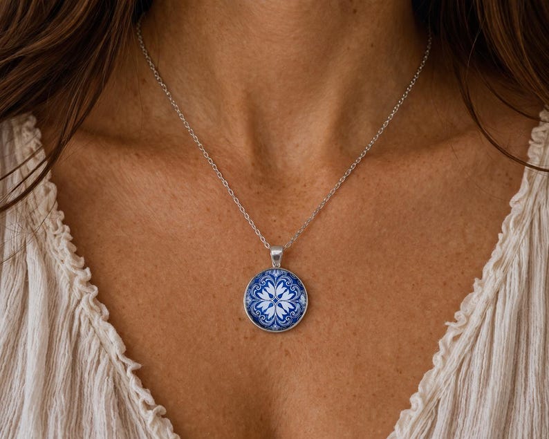 Puede incluir: Un collar de tono plateado con un colgante circular que presenta un patr&oacute;n floral en azul y blanco. El colgante cuelga de una delicada cadena de tono plateado que descansa sobre el pecho. El dise&ntilde;o muestra un motivo sim&eacute;trico y complejo.