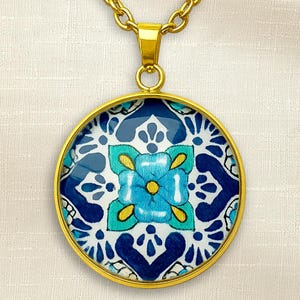 May include: A round pendant necklace featuring a gold-toned metal frame and a central circular insert. The insert displays a vibrant pattern of dark blue, light blue, and turquoise floral motifs with yellow accents on a white background.