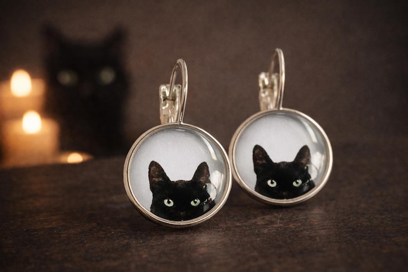 May include: A pair of round silver-toned drop earrings featuring a glass cabochon insert. Each earring displays a detailed photographic print of a black cat with bright green eyes against a white background, suspended from a silver-toned leverback.