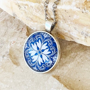 Puede incluir: Un colgante redondo de plata con un patr&oacute;n floral azul y blanco bajo un cabuj&oacute;n de cristal. El colgante est&aacute; unido a una fina cadena de plata, mostrando intrincados detalles de remolinos y un dise&ntilde;o sim&eacute;trico de p&eacute;talos en el centro.