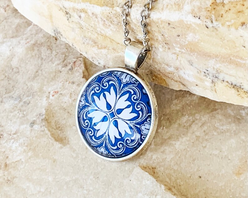 Puede incluir: Un colgante redondo de plata con un patr&oacute;n floral azul y blanco bajo un cabuj&oacute;n de cristal. El colgante est&aacute; unido a una fina cadena de plata, mostrando intrincados detalles de remolinos y un dise&ntilde;o sim&eacute;trico de p&eacute;talos en el centro.