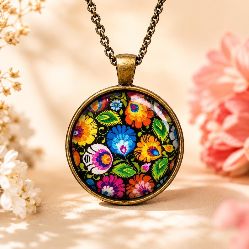 Puede incluir: Un collar con colgante redondo que presenta un engaste de metal en tono bronce y un caboch&oacute;n de vidrio. El inserto muestra un patr&oacute;n floral vibrante y multicolor con p&eacute;talos rojos, amarillos, azules y verdes sobre fondo negro.