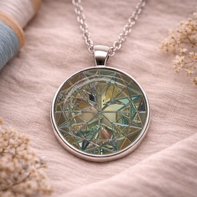 May include: A silver-toned pendant necklace featuring a circular charm with a complex, multi-faceted geometric pattern in shades of gold, teal, and silver. The pendant hangs from a delicate silver-colored metal chain with small, uniform links.