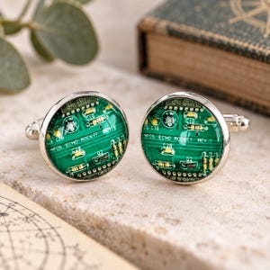 May include: A pair of round silver-toned cufflinks featuring a green circuit board pattern under a glass dome. The circuit board design includes yellow resistors and white text that reads MFOS ECHO ROCKIT REV 1, set within a polished metal frame.