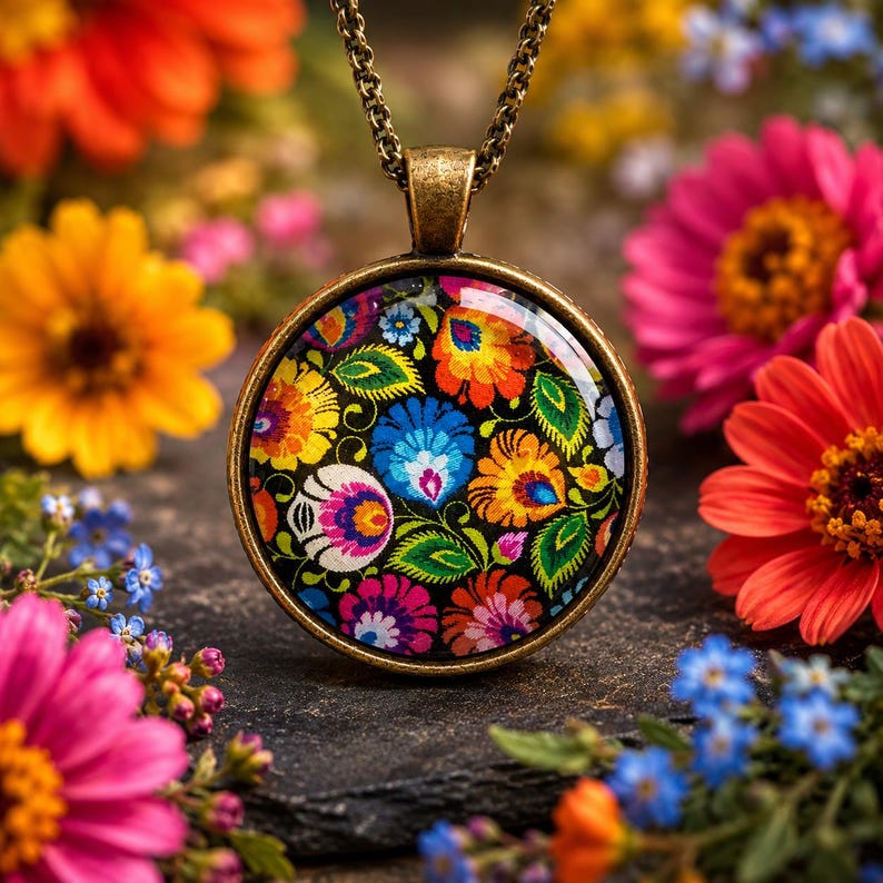 Puede incluir: Un collar con colgante redondo que presenta un vibrante patr&oacute;n floral con p&eacute;talos naranjas, azules, rosas y verdes sobre un fondo negro, encerrado en un marco de metal color bronce y suspendido de una cadena a juego.