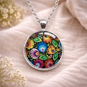Puede incluir: Un collar con colgante plateado que presenta un cabochon circular de cristal. El inserto muestra un vibrante patr&oacute;n floral multicolor con p&eacute;talos y hojas en rojo, naranja, amarillo, azul y verde sobre un fondo negro s&oacute;lido.