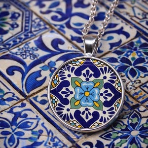 May include: A round silver pendant necklace featuring a colorful tile-inspired pattern. The design displays a central light blue flower with a yellow center, surrounded by dark blue, green, and white geometric and floral motifs on a circular metal base.