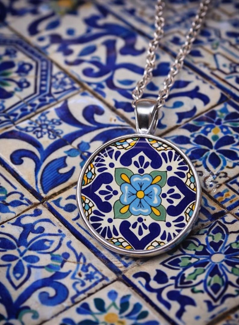 May include: A round silver pendant necklace featuring a colorful tile-inspired pattern. The design displays a central light blue flower with a yellow center, surrounded by dark blue, green, and white geometric and floral motifs on a circular metal base.