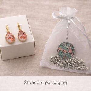 May include: A pair of gold-toned teardrop earrings with a pink and white floral pattern resting on a white box, alongside a silver-toned circular pendant necklace with a dark green and pink floral design inside a sheer white drawstring pouch.
