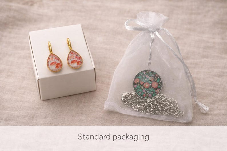 May include: A pair of gold-toned teardrop earrings with a pink and white floral pattern resting on a white box, alongside a silver-toned circular pendant necklace with a dark green and pink floral design inside a sheer white drawstring pouch.