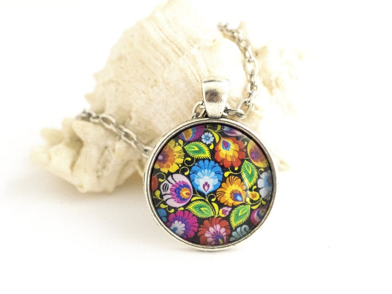 Puede incluir: Un collar con colgante redondo de plata que presenta un vibrante patr&oacute;n floral multicolor sobre un fondo negro. El dise&ntilde;o incluye intrincados motivos de flores y hojas en rojo, azul, amarillo y verde dentro de un marco de metal plateado pulido.