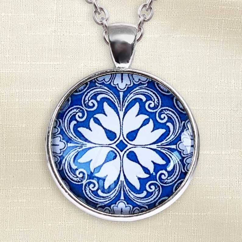 Puede incluir: Un colgante circular de plata con un vibrante patr&oacute;n floral en azul y blanco. El dise&ntilde;o consiste en p&eacute;talos blancos sim&eacute;tricos dispuestos en forma de cruz sobre un fondo azul intenso con detalles intrincados de volutas blancas.