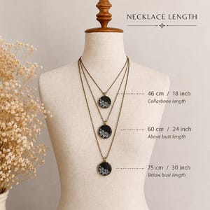 Puede incluir: Tres collares de tono bronce con colgantes redondos negros con patrones de encaje floral blanco. Las longitudes indicadas son 46 cm (clav&iacute;cula), 60 cm (sobre el busto) y 75 cm (bajo el busto) en un maniqu&iacute;.