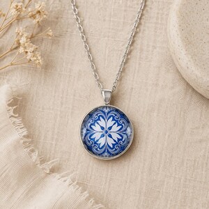 Puede incluir: Un collar de metal plateado con un colgante circular que presenta un patr&oacute;n floral en azul y blanco. El colgante cuelga de una cadena plateada a juego. El dise&ntilde;o muestra un motivo sim&eacute;trico e intrincado que recuerda a los azulejos.
