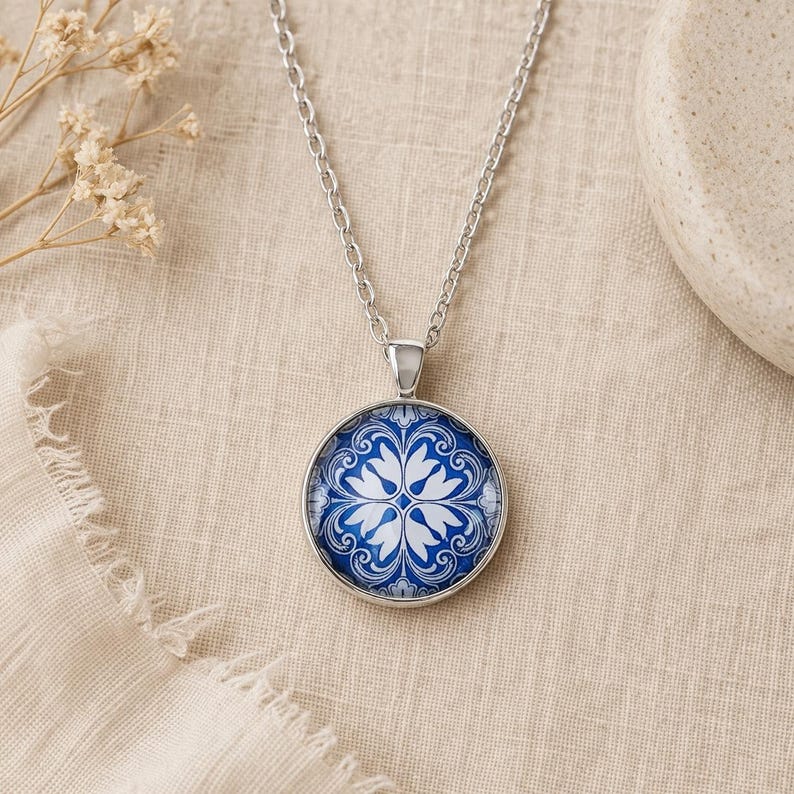 Puede incluir: Un collar de metal plateado con un colgante circular que presenta un patr&oacute;n floral en azul y blanco. El colgante cuelga de una cadena plateada a juego. El dise&ntilde;o muestra un motivo sim&eacute;trico e intrincado que recuerda a los azulejos.