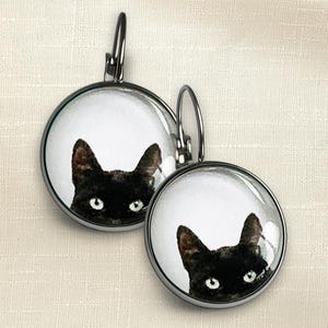 May include: A pair of round leverback earrings featuring a black cat illustration on a white background. The earrings have a dark metal frame and a glass cabochon dome that magnifies the cat's face, showing its ears and bright eyes clearly.
