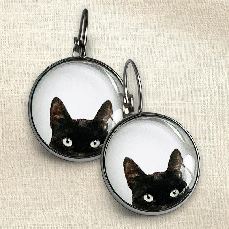 May include: A pair of round leverback earrings featuring a black cat illustration on a white background. The earrings have a dark metal frame and a glass cabochon dome that magnifies the cat's face, showing its ears and bright eyes clearly.
