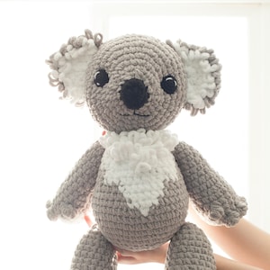 Puede incluir: Un koala de peluche gris tejido a crochet con orejas blancas de crochet y un parche de pecho blanco de crochet. El koala está sentado con los brazos extendidos.