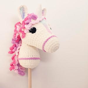 May include: A white crocheted unicorn rocking horse with pink and purple yarn mane and tail. The unicorn has a white horn and black eyes. The rocking horse is attached to a wooden stick.