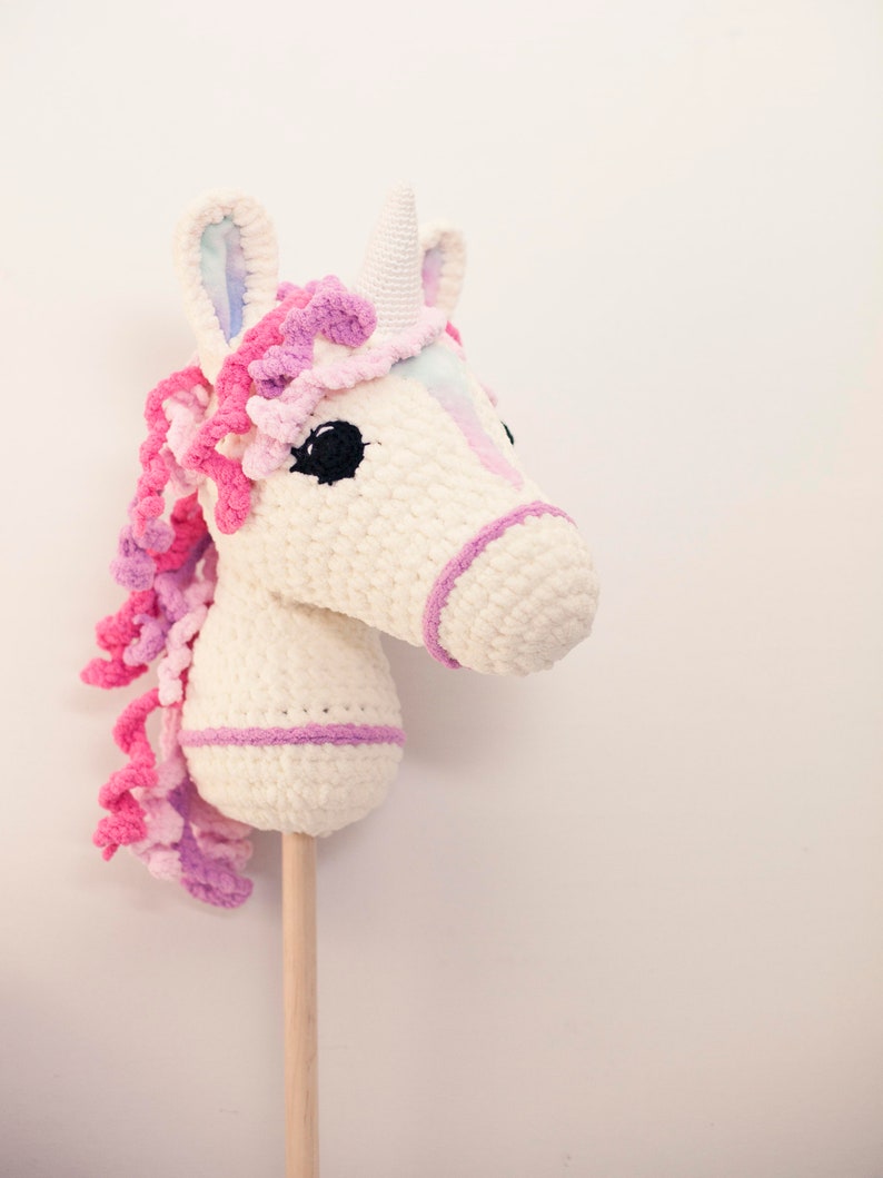 May include: A white crocheted unicorn rocking horse with pink and purple yarn mane and tail. The unicorn has a white horn and black eyes. The rocking horse is attached to a wooden stick.