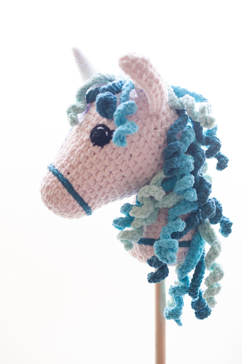 May include: A pink and blue crocheted unicorn head on a wooden stick. The unicorn has a blue and green mane and a white horn.