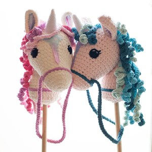 May include: Two crocheted unicorn stick horses, one pink with blue mane and the other white with pink mane, are standing side by side. Both have wooden sticks and reins.
