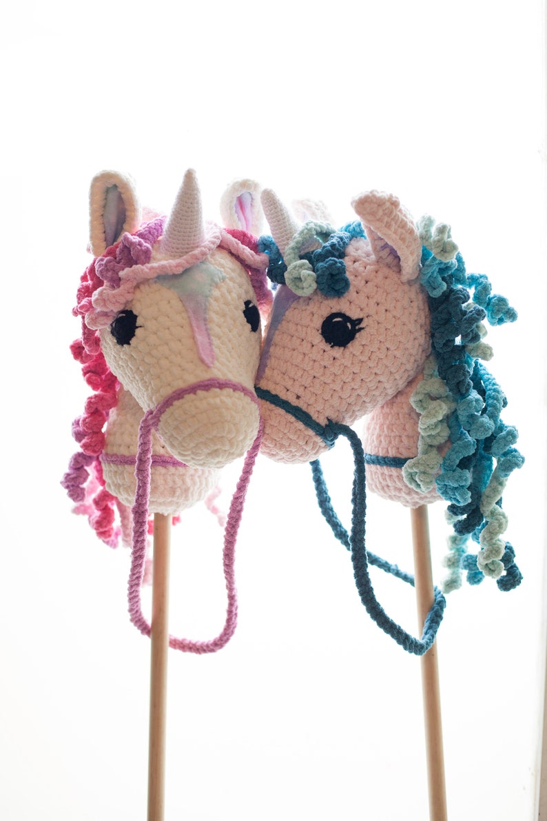 May include: Two crocheted unicorn stick horses, one pink with blue mane and the other white with pink mane, are standing side by side. Both have wooden sticks and reins.