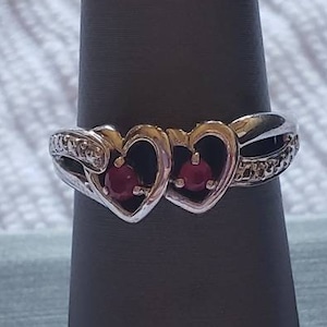 May include: A silver ring with two intertwined hearts, each set with a ruby gemstone. The ring is accented with small diamonds.