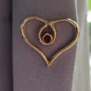 Gold plated heart lapel pin. This simple heart pin has a small glass red stone in the center of the heart. The pin is gold plated.