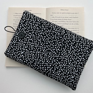 May include: A black and white polka dot book cover with a button closure and a black elastic strap. The cover is lying on top of an open book.