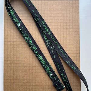 May include: Black lanyard with a green binary code pattern. The lanyard has a silver-colored metal clasp and is lying on a brown grid-lined paper. The lanyard is made of a soft, durable fabric.