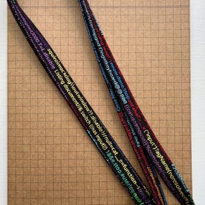 Può includere: Un laccio nero con un moschettone argentato. Il laccio ha del testo colorato stampato su di esso, inclusi frammenti di codice e le parole "dog woof", "meow", "done", "stop", "false", "input", "TagName", "noresolve", "function", "string", "userlog", "node", "type", "cat", "hashtag", "algorithm", "Lorem", "ipsum", "logged", "oft", "close", "filter", "window", "close", "car", "reload", "upload", "download", "loosquireimackdog", "outwindow", "disable", "null", "cat", "e", "function", "string", "str", "kedogout", "T", "L", "disable", "Listing", "document", "switch", "has", "input", "1", "e", "stop", "userlog", "node", "type", "39", "home", "resolve", "p".