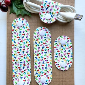 May include: Four white cable organizers with a Christmas light pattern. The organizers have white buttons and are on a brown grid background.
