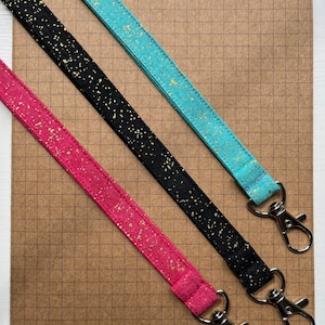 May include: Three fabric lanyards with gold glitter on a brown grid background. The lanyards are black, pink, and turquoise. Each lanyard has a silver swivel clasp.
