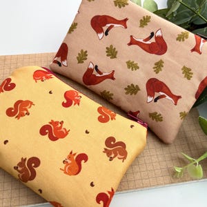 May include: Two rectangular zipped pouches. One is beige with a pattern of red foxes and green leaves. The other is yellow with a pattern of orange and brown squirrels. Both pouches are on a notebook.